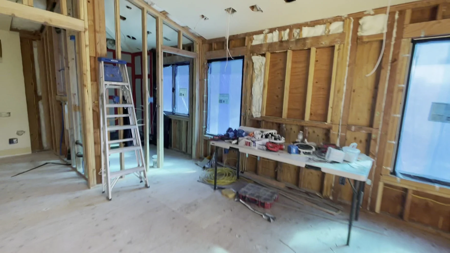 Interior at framing stage with reframed rectangular openings, glazed units and door assemblies waiting on site to go in, and exposed studs and ceiling joists