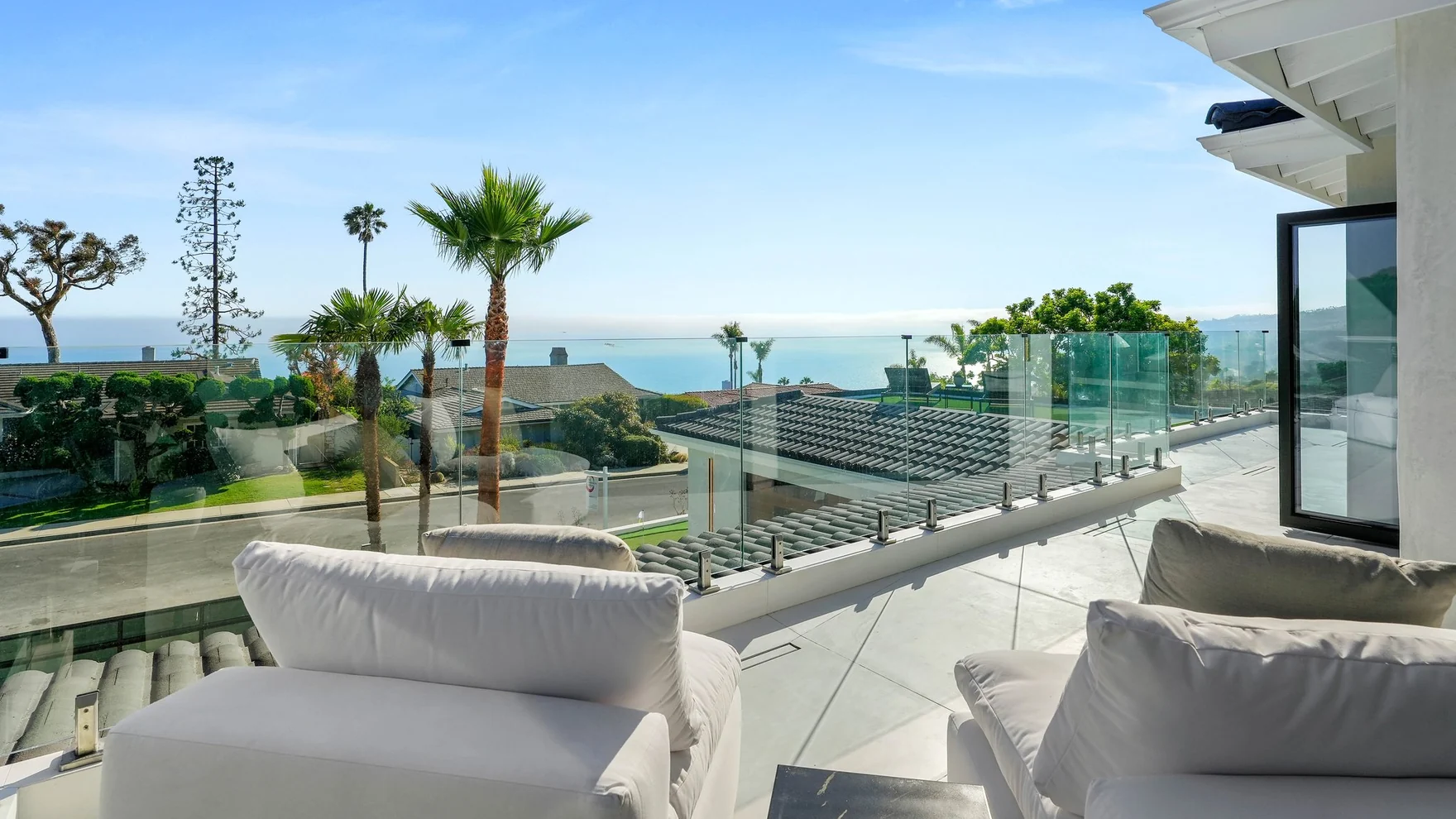The same deck rebuilt — a glass balustrade with no posts, pale seating, palm heads and nothing but horizon past them