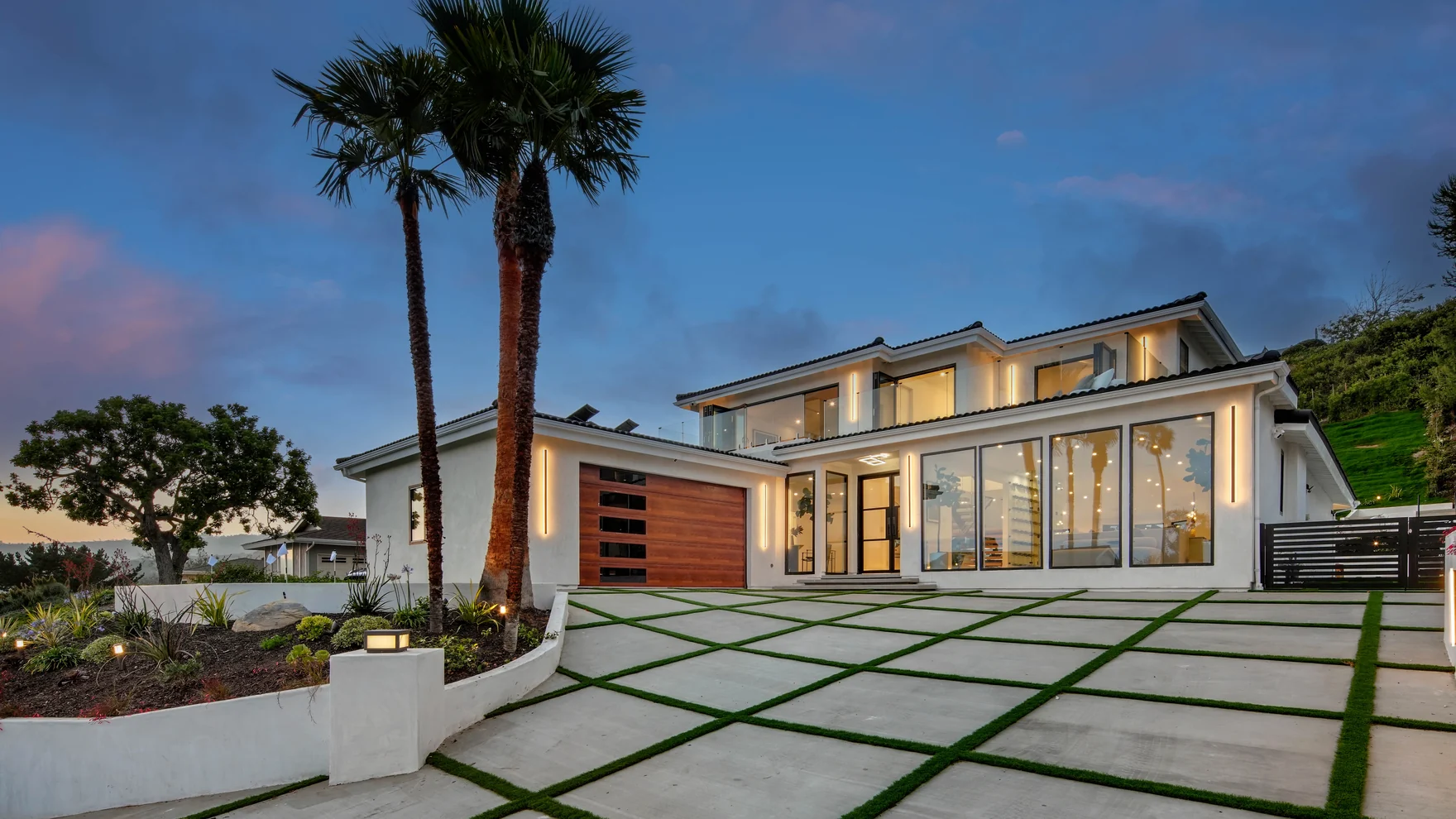 Rebuilt modern street elevation at dusk with a low horizontal roofline, glowing glass, a slatted timber panel, palms and a drive of concrete bands set in grive
