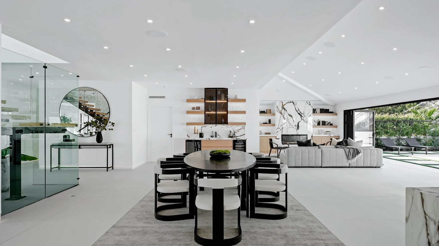 The rebuilt level — a round black table, pale oak shelves cantilevered off white plaster, stone slab, and glazing the full width of the room