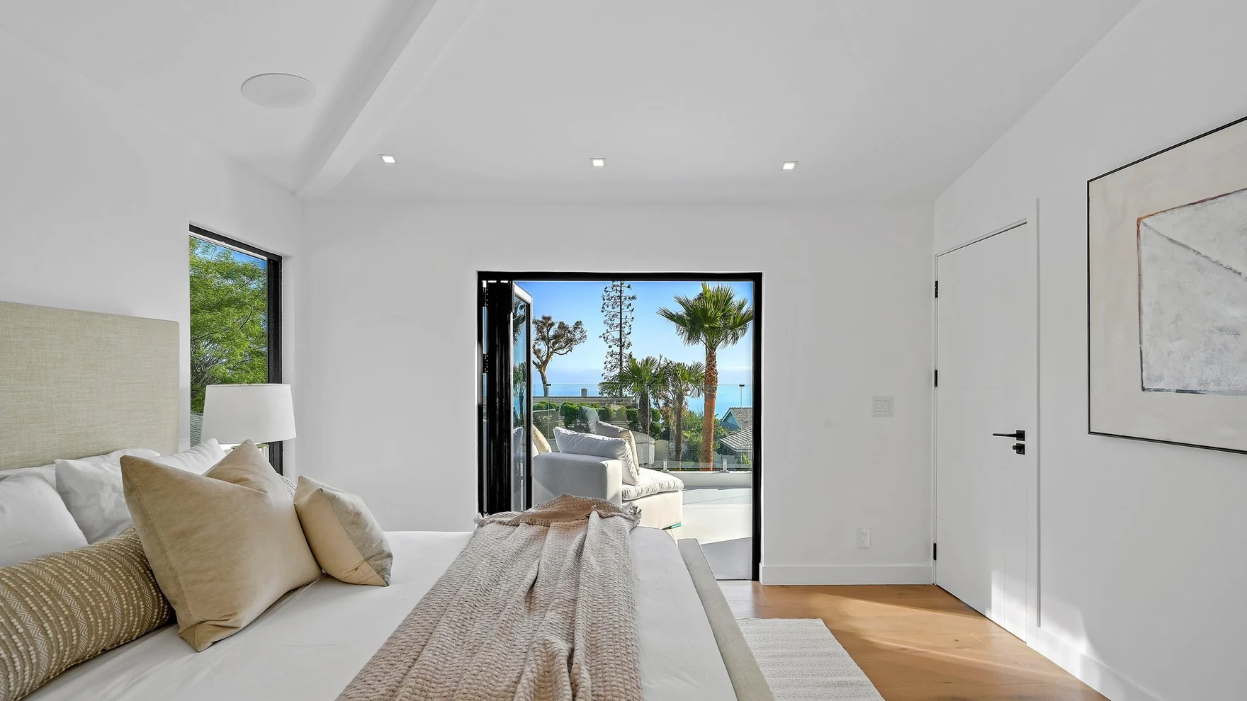 Guest bedroom with a vaulted white ceiling, white oak floor and a folding glass door open to a terrace with the ocean beyond
