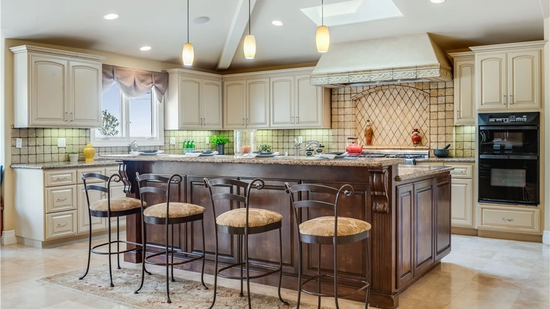 Kitchen as found — cream cabinet fronts, speckled stone tops, small square splash tiles and iron stools on a stone-tiled floor
