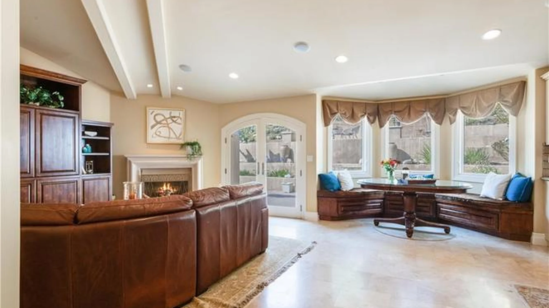 Original beige family room with a brown leather sectional, arched windows behind fabric valances, stained-wood built-ins and a patterned rug