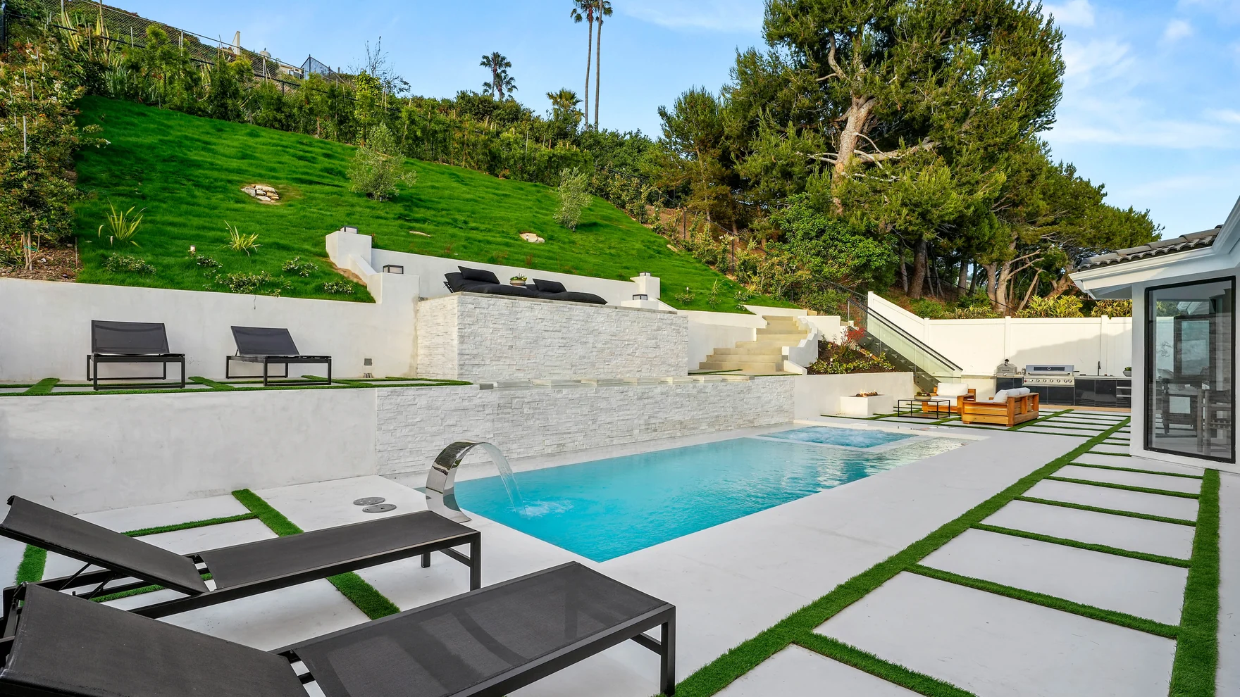 New rectangular pool with a sun shelf, black-frame chaise lounges and concrete-and-turf grid decking against the green slope