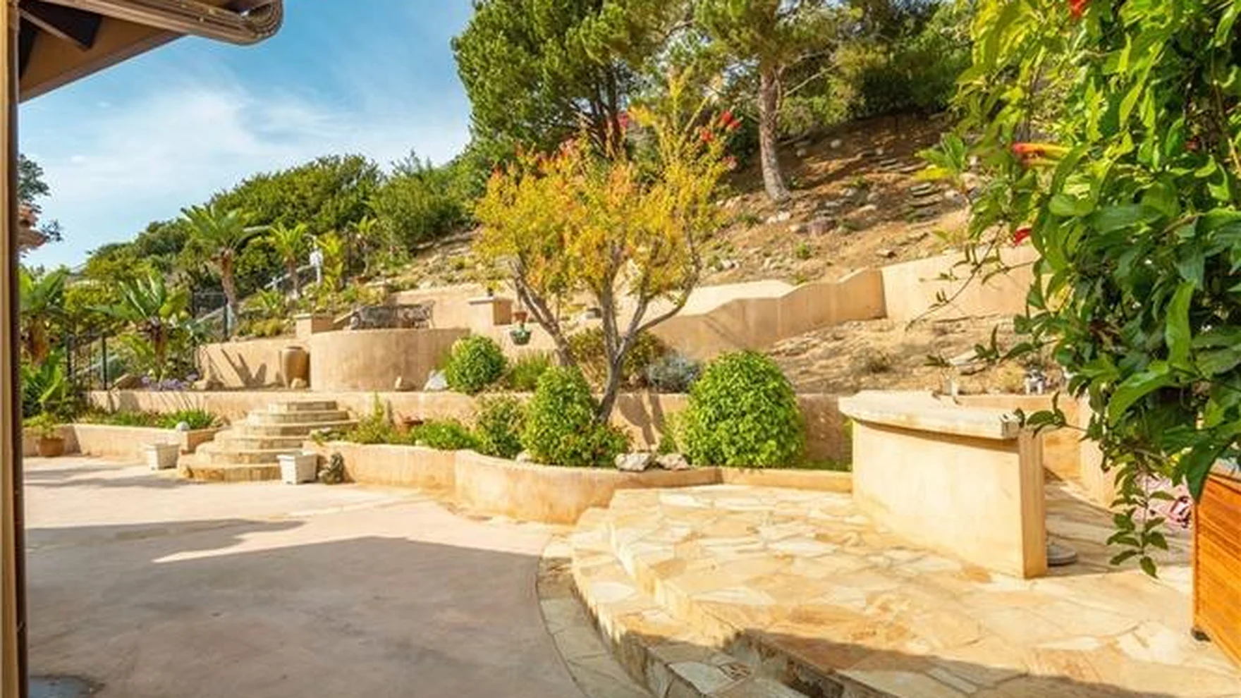 Original rear yard on a bare slope with stone retaining walls, dirt and patchy grass, mature trees stepping up the hillside
