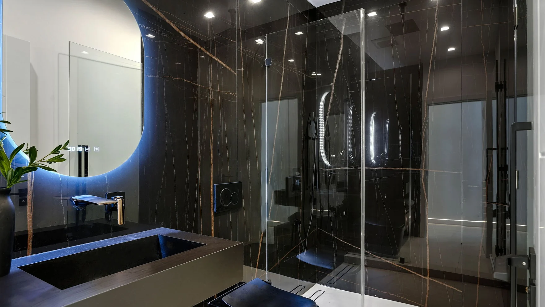 Bathing room lined in dark veined stone, a glass screen with no frame, darkened metal fittings and a tub standing clear of the wall