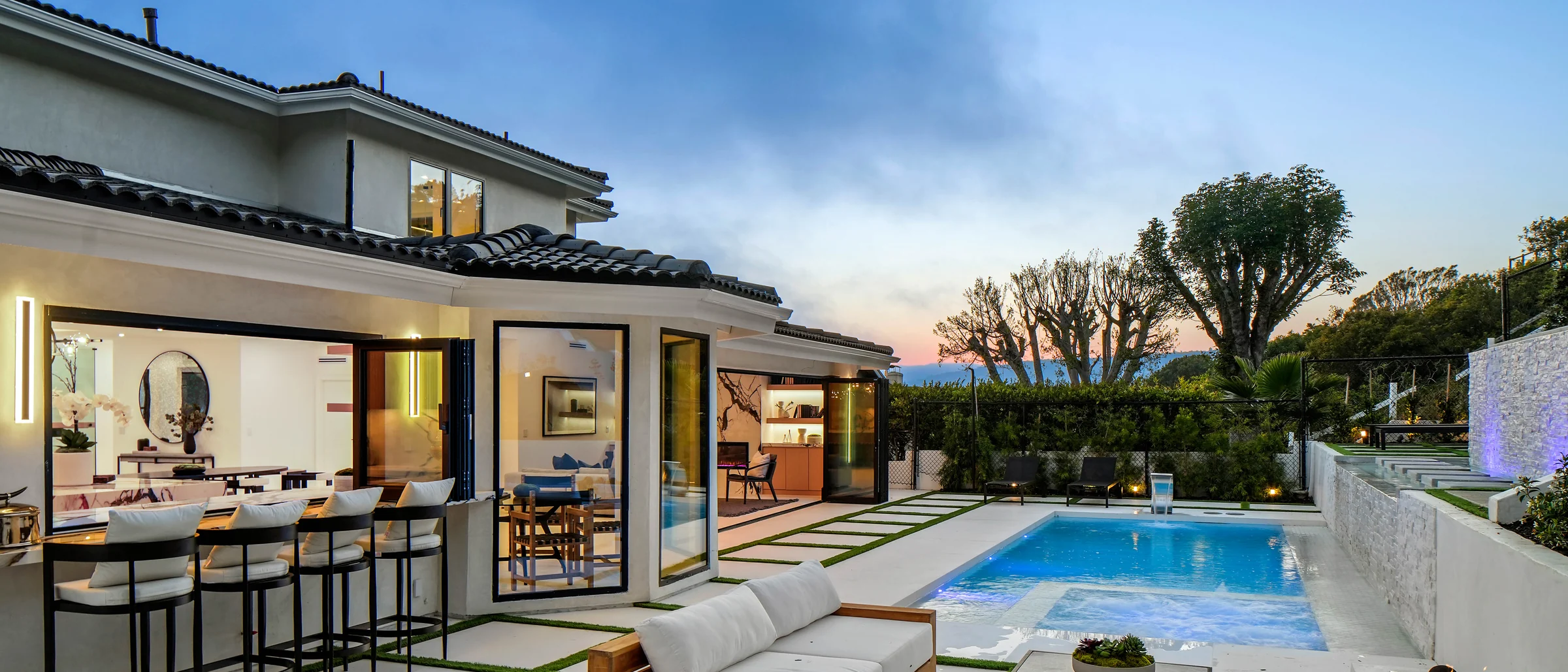 Rear elevation of the rebuilt house at twilight: full-height glass walls open, the lit interior reading through to the pool